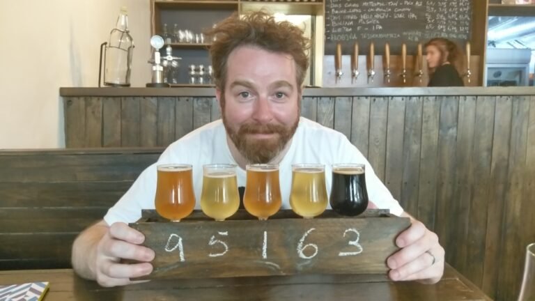 A mid-30s bearded white guy sitting in a craft beer bar in front of a 5 glass beer flight in a wooden box.